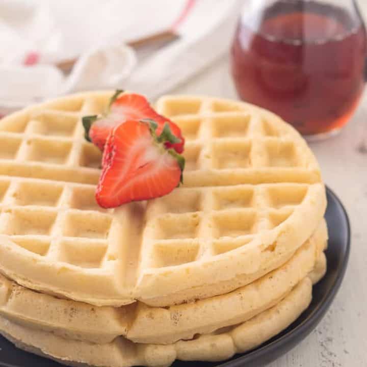 stack of three gluten free waffles with a sliced strawberry on top and pitcher of maple syrup