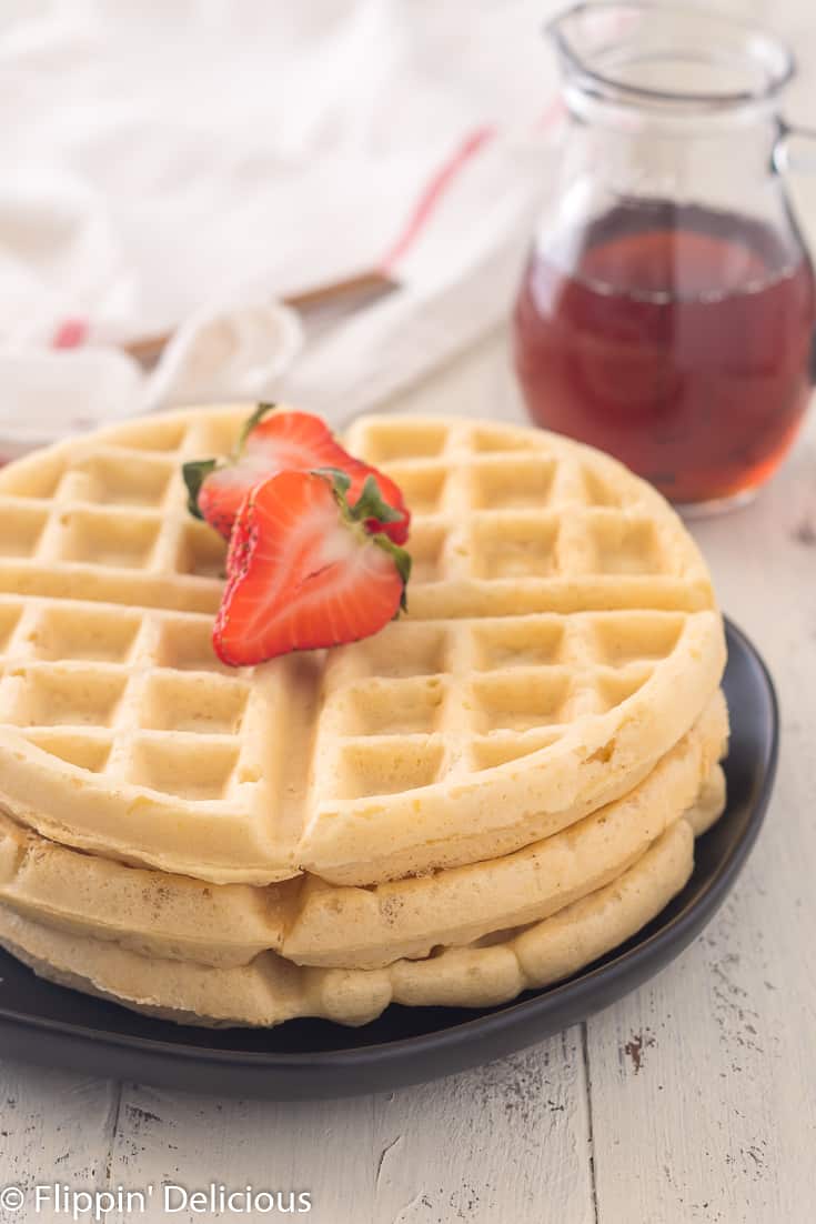 stack of three gluten free waffles with a sliced strawberry on top and pitcher of maple syrup