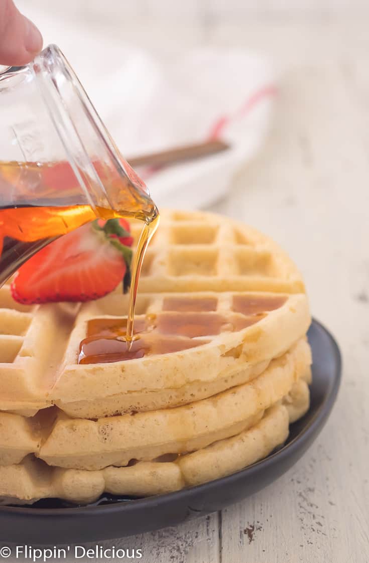 pouring maple syrup on stack of three gluten free Belgian waffles with sliced strawberry