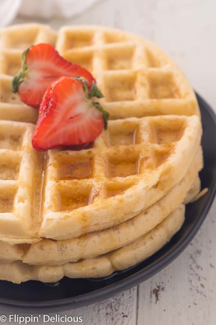 stack of three gluten free waffles on a plate with a halved strawberry and maple syrup