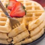 fork taking a bite out of stack of three gluten free waffles on plate with maple syrup and sliced strawberry