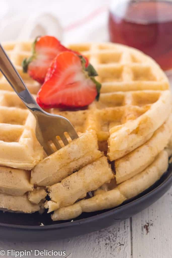 fork taking a bite out of stack of three gluten free waffles on plate with maple syrup and sliced strawberry