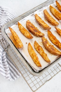 oven baked potato wedges on baking wheet lined with foil on top of a cooling rack with a dishtowel in the foreground
