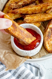 fingers holding a seasoned baked potato wedge and dipping into a white bowl with ketchup