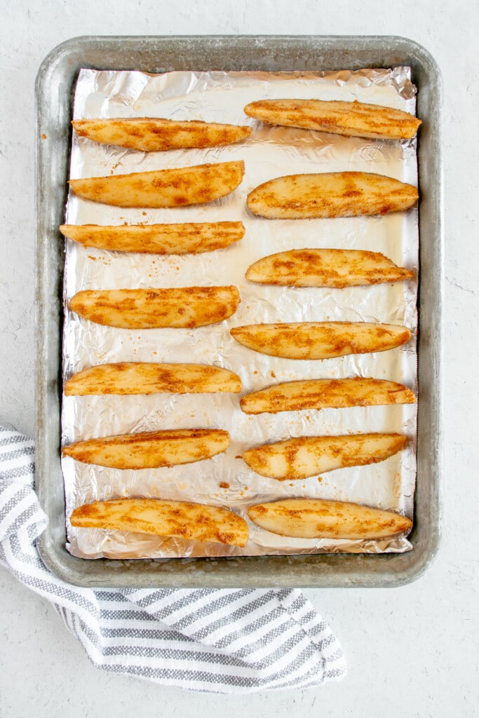 Overhead photo of pan lined with foil with potato wedges seasoned with oil, cajun seasoning, gluten free flour, salt, and pepper ready to bake beside a striped dish towel 