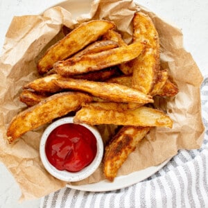 overhead view of seasoned potato wedges oven roasted to perfection on a plate lined with brown paper and a small bowl of ketchup on a marble table top with a striped dish towel