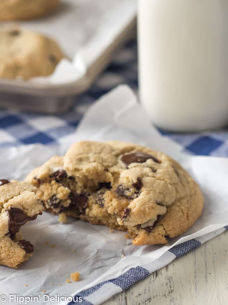 A moist gluten free chocolate chip cookie broken in half on a piece of white parchment paper on a blue checked napkin