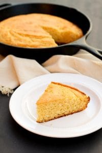 gluten free corn bread wedge on a white plate with skillet of gluten free honey cornbread in the background