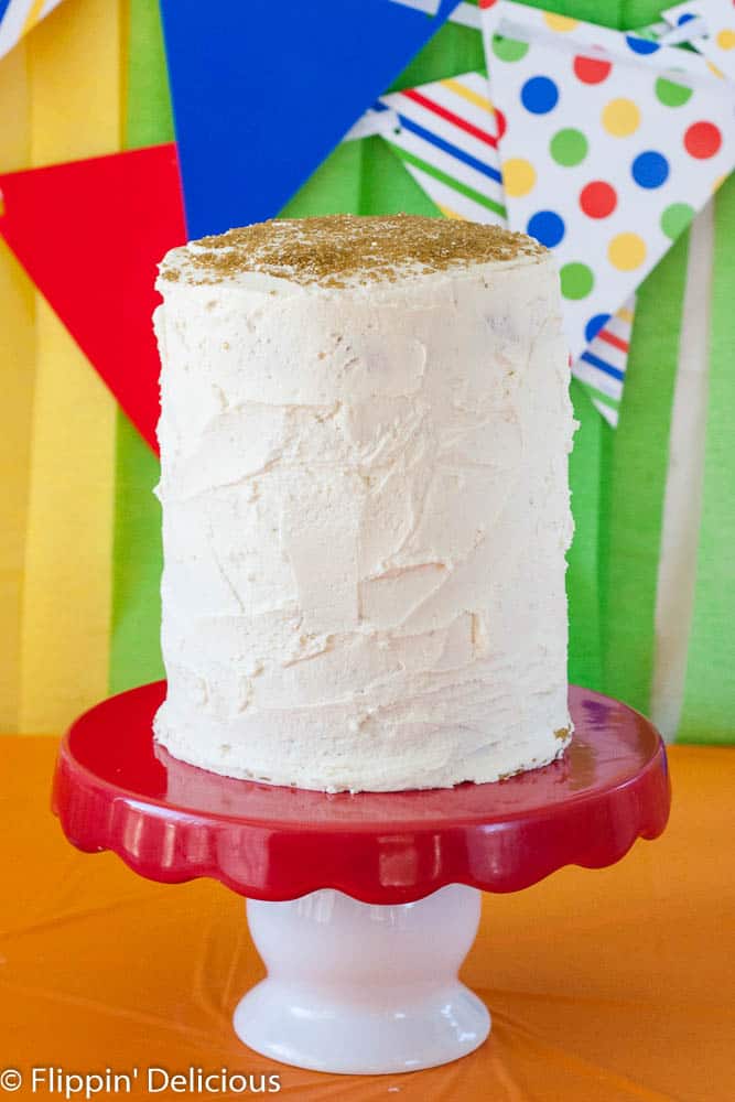 a gluten free white frosted 6 layer 6 inch cake on a red and wite cake stand, with rainbow streamers and banners in the background