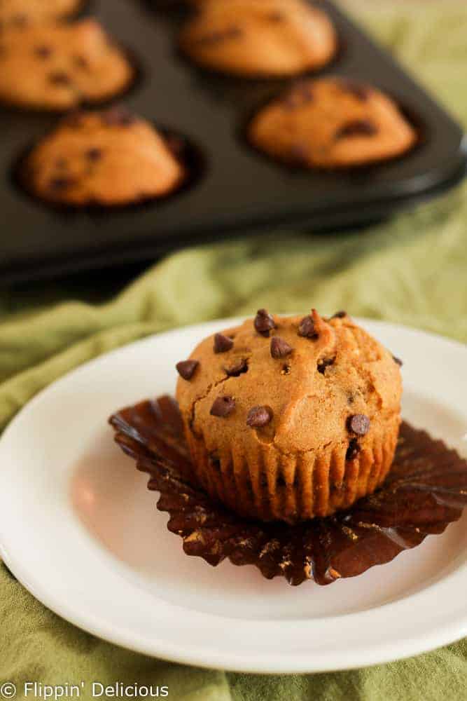 close up of unwrapped gluten free pumpkin muffin with chocolate chips on a brown muffin liner and a white plate, on top of a green table cloth with gluten free pumpkin muffins in the background in a muffin tin