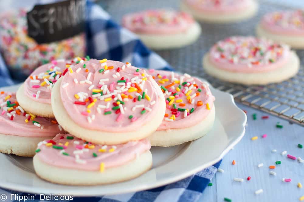 white plate with round gluten free sugar cookies with pink frosting and rainbow sprinkles with more sprinkles and cookies in the background on a cooling rack