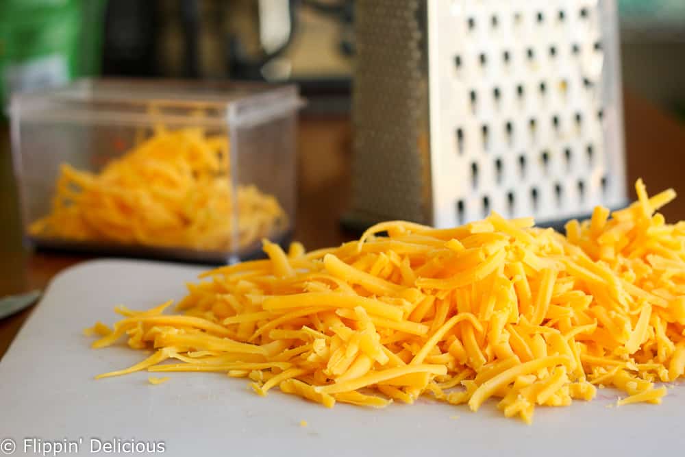 freshly grated cheddar cheese on a cutting board with a cheese grater in the background