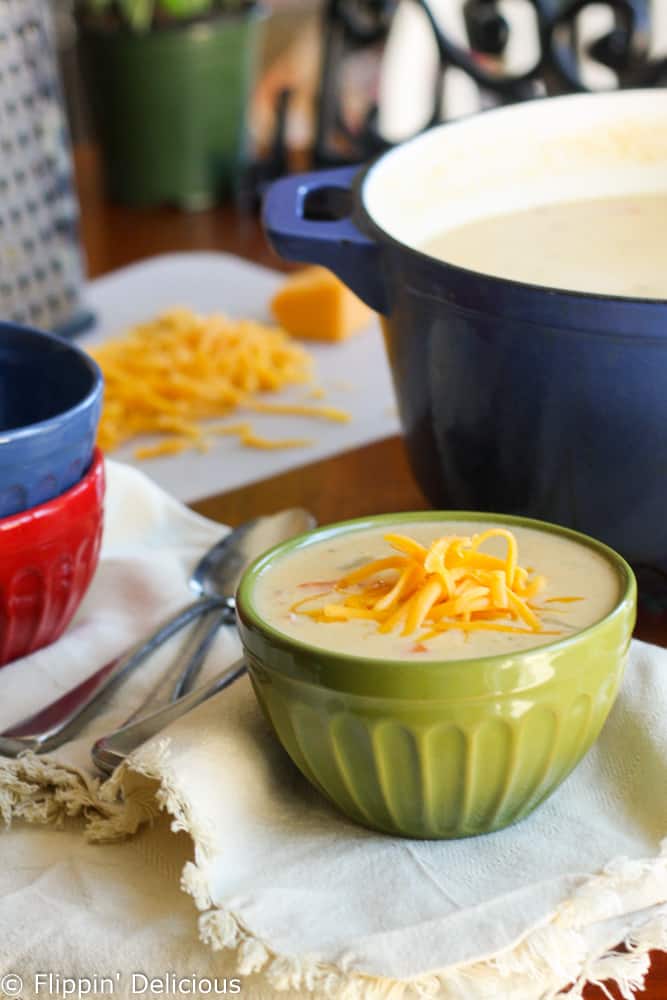 green bowl filled with gluten free cheddar cheese soup with ham topped with grated cheese, on a cloth napkin with spoons and a blue dutch oven in the backgroud