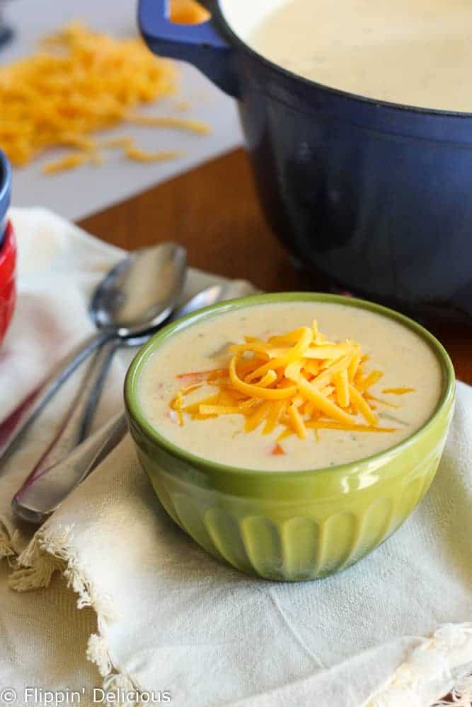 green bowl filled with gluten free ham and cheese soup topped with grated cheese, with spoons and a blue dutch oven in the backgroud