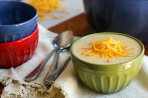 green bowl filled with cheddar cheese with ham sprinkled with grated cheddar cheese on a wooden table with a stack of bowls and spoons in the background