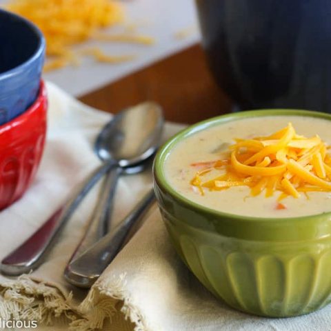 green bowl filled with cheddar cheese with ham sprinkled with grated cheddar cheese on a wooden table with a stack of bowls and spoons in the background