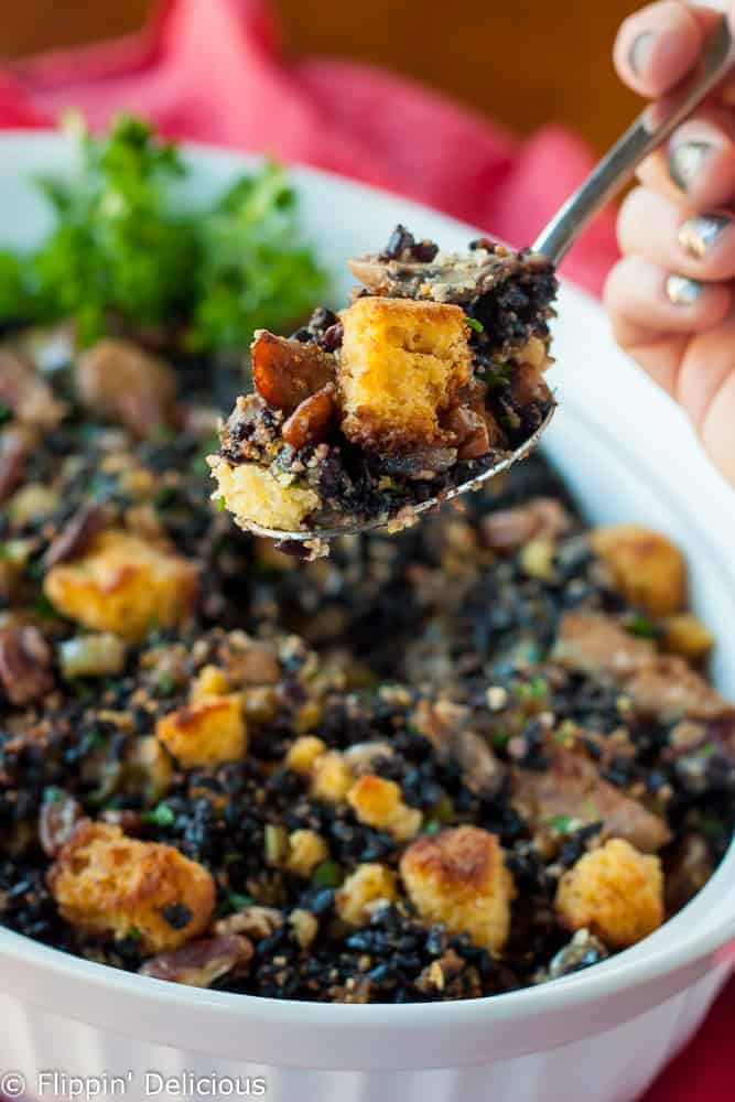 spoon with gluten free cornbread dressing with pecans, wild rice, and andouille sausage with a casserole dish full of gluten free cornbread dressing in the background