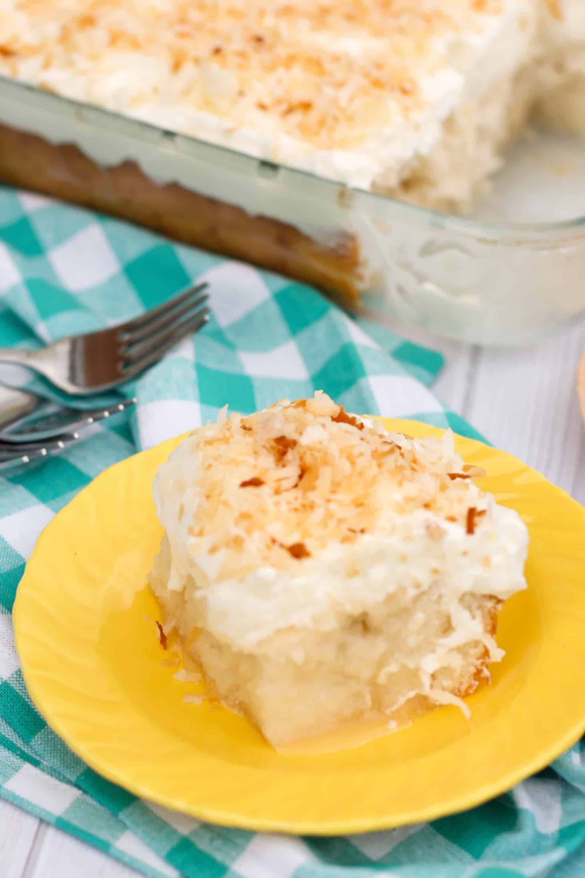 square slice of coconut poke cake sprinkled with toasted coconut on a yellow plate with a teal plaid napkin in the background