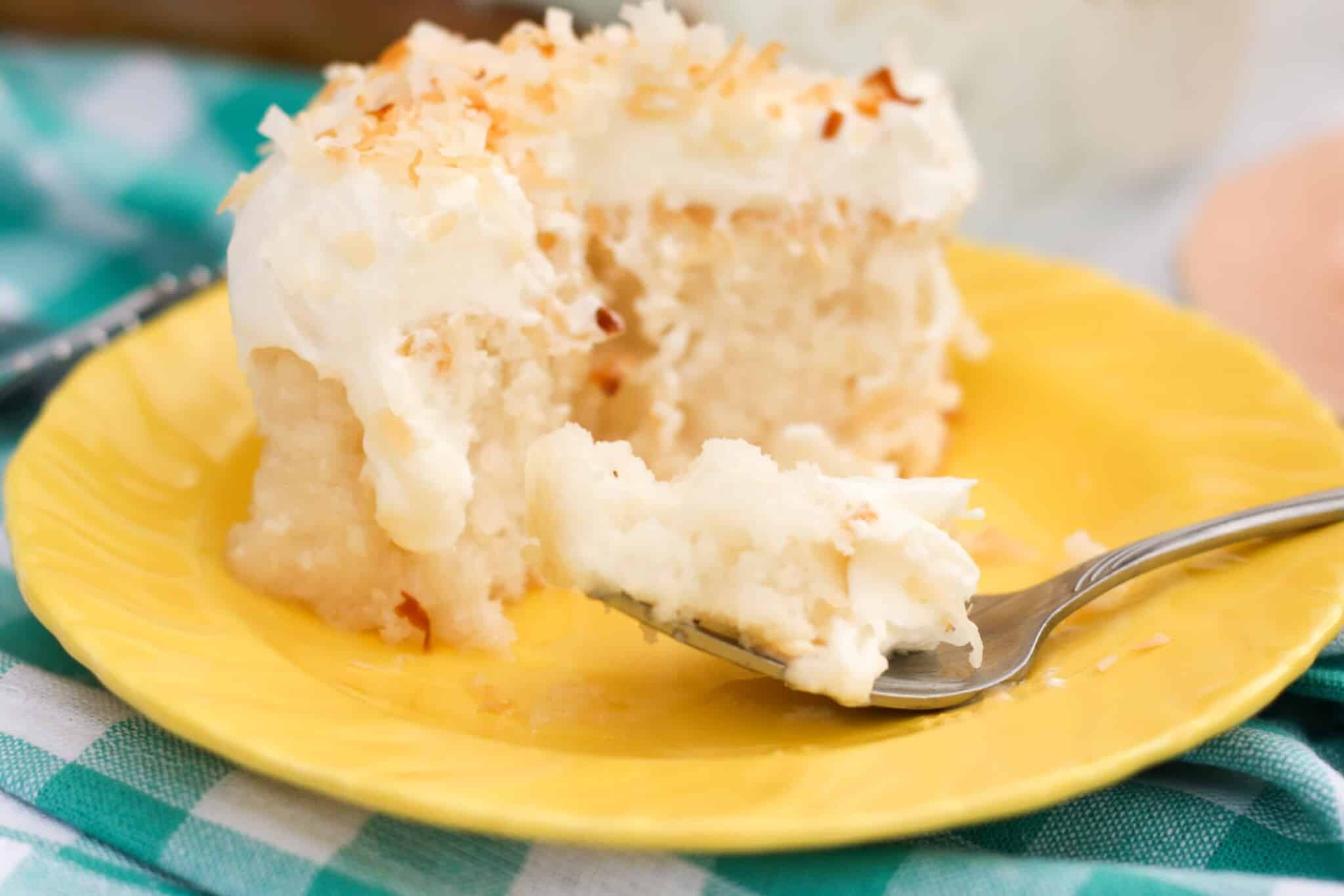 slice of dairy free gluten free coconut poke cake with a fork taking a bite out of the coconut poke cake, on a yellow plate.