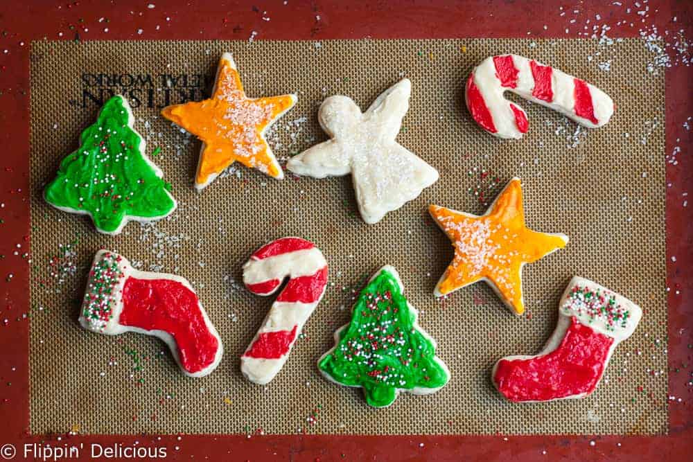 gluten free vegan sugar cookie cut outs in christmas and holiday shapes, frosted and sprinkled on a silpat baking mat
