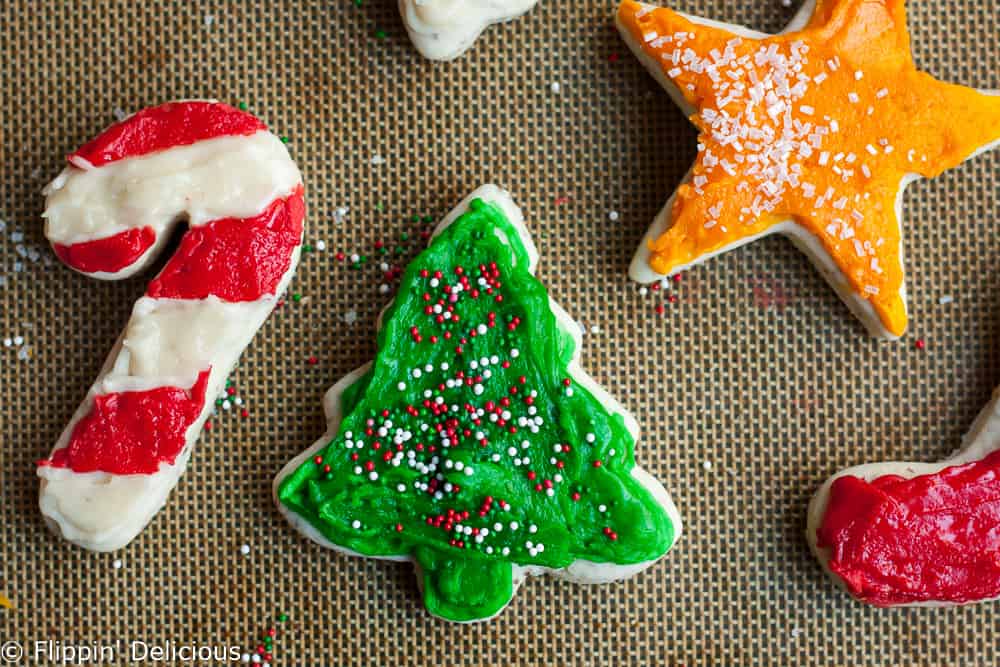 vegan sugar cookie cut outs in the shapes of christmas trees, holiday candy canes, and stars frosted ith red, green, and gold frosting and covered ith sprinkles on a silpat baking mat