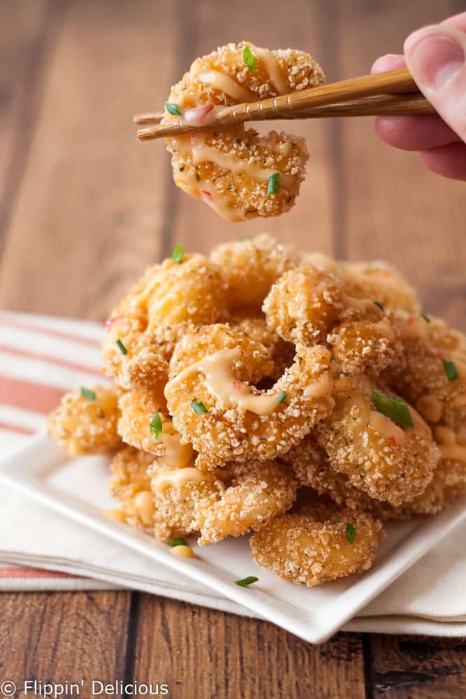 a pile of gluten free fried breaded shrimp drizzled in bang bang sauce, ith a pair of chopsticks holding a gluten free ban bang shrimp over the plate