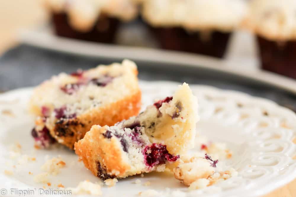 piece of gluten free blueberry muffin on a white plate, beside a half of a gluten free blueberry muffin with streusel crumb topping with a platter of muffins in the background