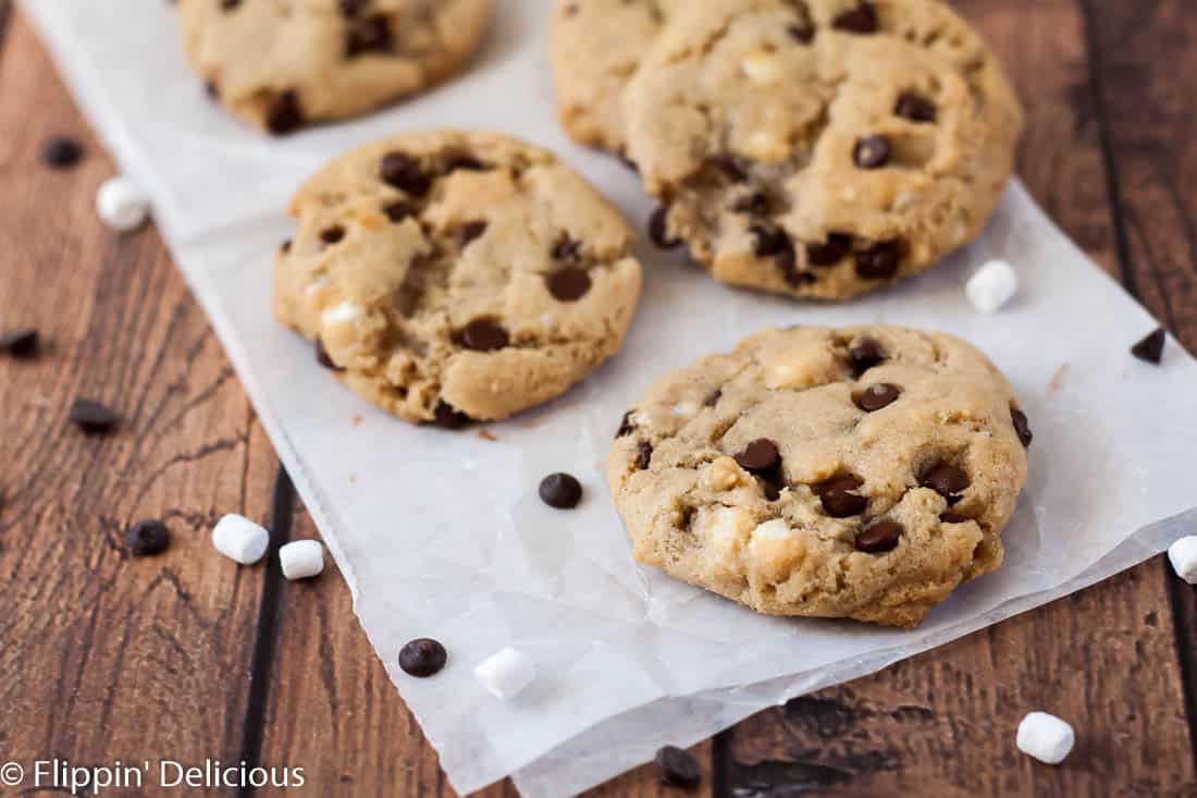 several gluten free dairy free chocolate chip cookies with mini chocolate chips ad dehydrated mini marshmallows on a piece of parchment paper on a wooden background