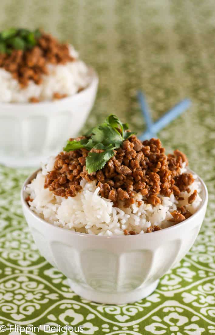 white bowl filled with rice and ground turkey seasoned with tamari and brown sugar, garnished with cilantro , on a green and white table cloth with blue chopstick in the background