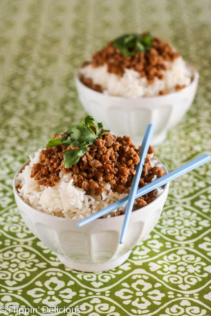 white bowl filled with rice and ground turkey seasoned with tamari and brown sugar, garnished with cilantro and blue chopsticks, on a green and white table cloth.