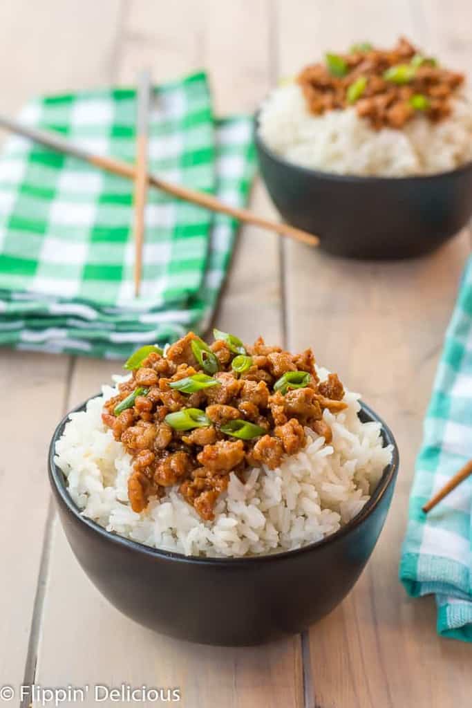 black matte bowl of rice topped with gluten free korean ground turkey garnished with green onions on a wooden table with another gluten free korean ground turkey and rice bowl in the background with teal and green napkins and wooden chopsticks