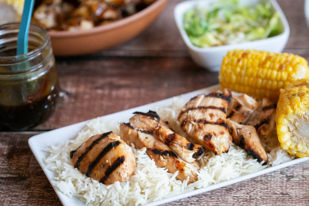 grilled gluten free teriyaki chicken on bed of rice on white plate with grilled corn on the cob