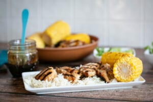 grilled gluten free teriyaki chicken on a white plate with rice and corn on a wooden table with homemade gluten free teriyaki sauce in a jar in the background