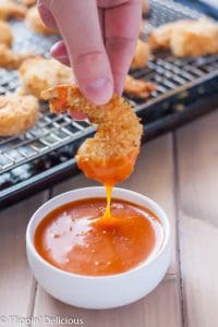 fingers holding gluten free coconut shrimp being dipped into orange colored sauce in a white bowl