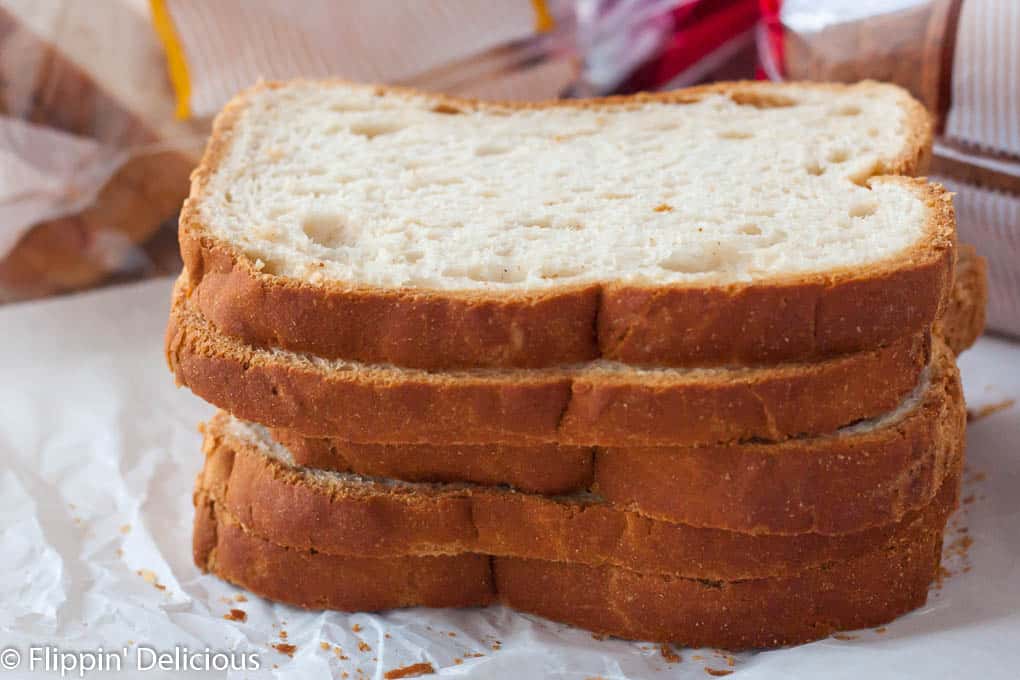 5 Scheiben glutenfreies Canyon Bakehouse Heritage Honey White Bread übereinander gestapelt, auf weißem Pergamentpapier mit Paketen von glutenfreiem Brot im Hintergrund