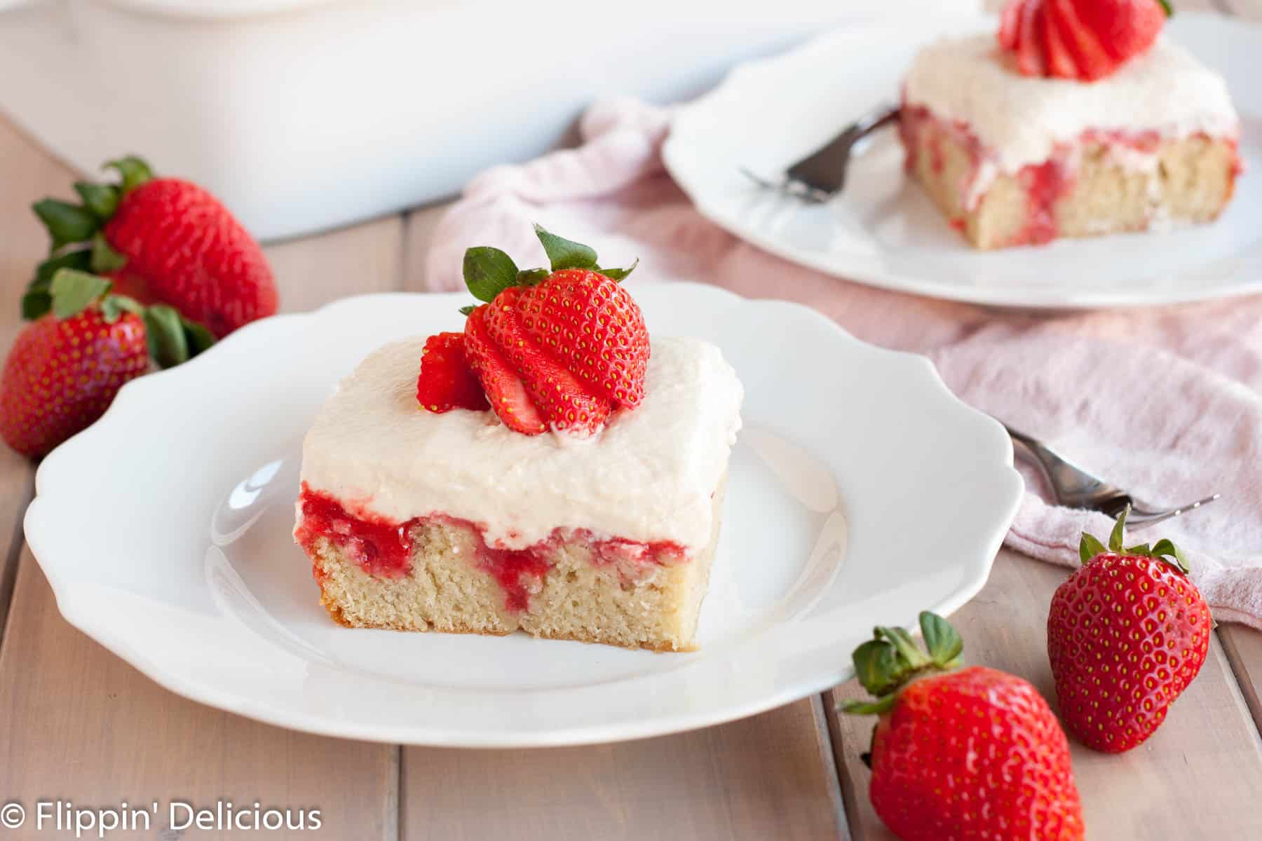 a slice of gluten free strawberry poke cake topped with a fresh strawberry on a white plate with scalloped edges on a light gray wooden table with fresh strawberries