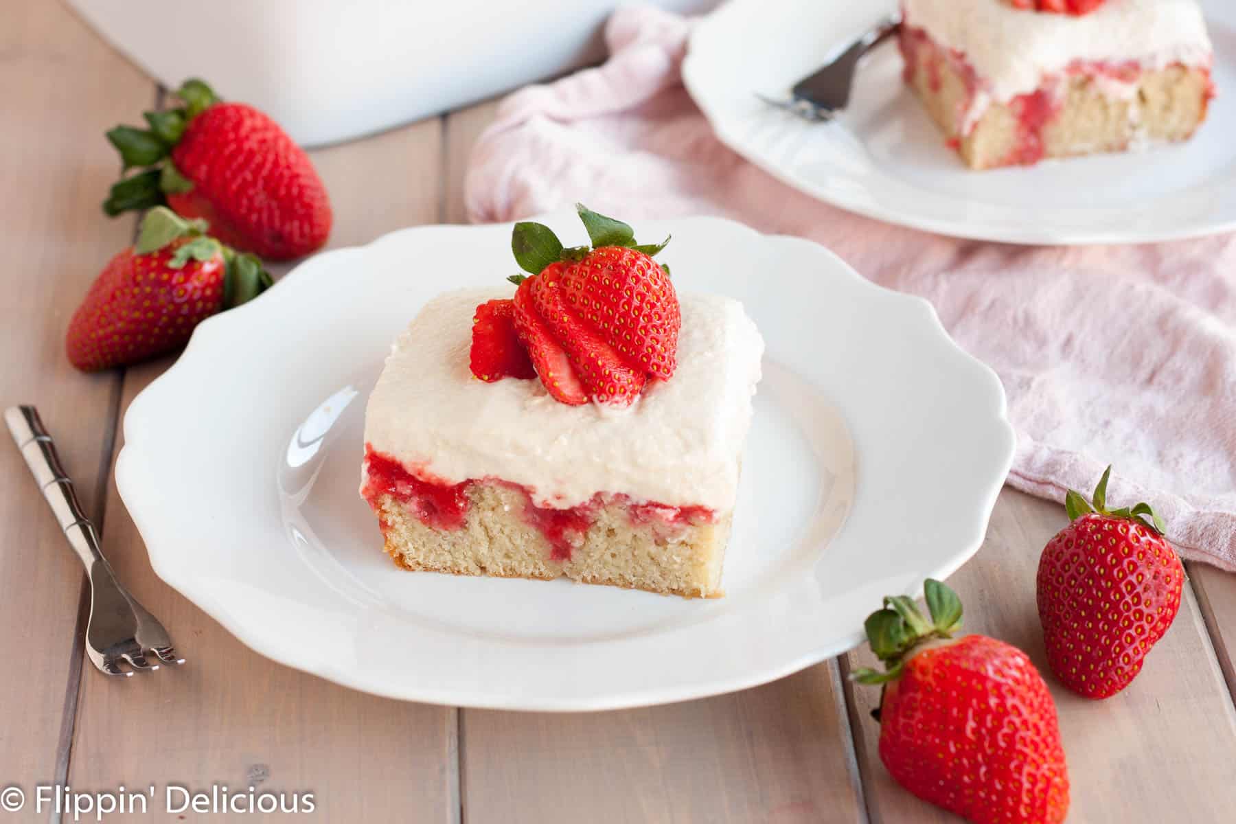 A slice of gluten free strawberry cake topped with fluffy pudding, strawberry sauce, and a fresh strawberry, sliced on top of a white plate on a light grey wood table with a small fork, fresh strawberries, and a light pink dish towel 