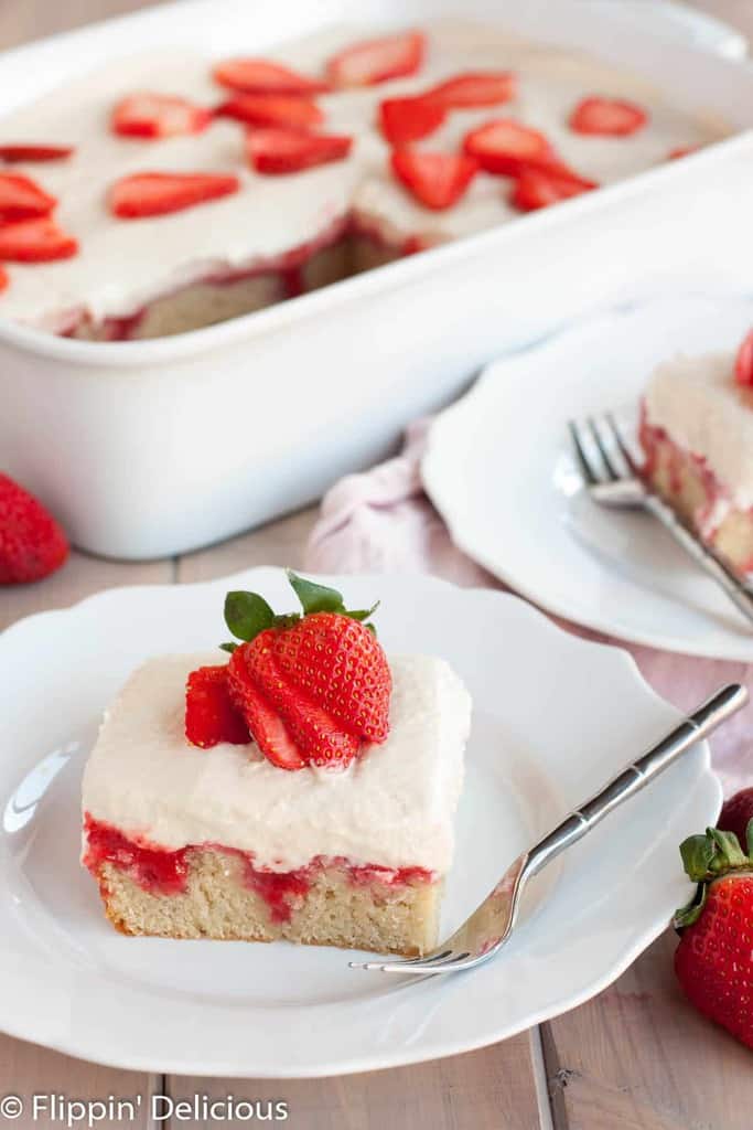 a slice of gluten free poke cake covered in homemade strawberry syrup and topped with a fluffy dairy free pudding and a sliced fresh strawberry, with a white pan with more gluten free strawberries and cream poke cake in the background