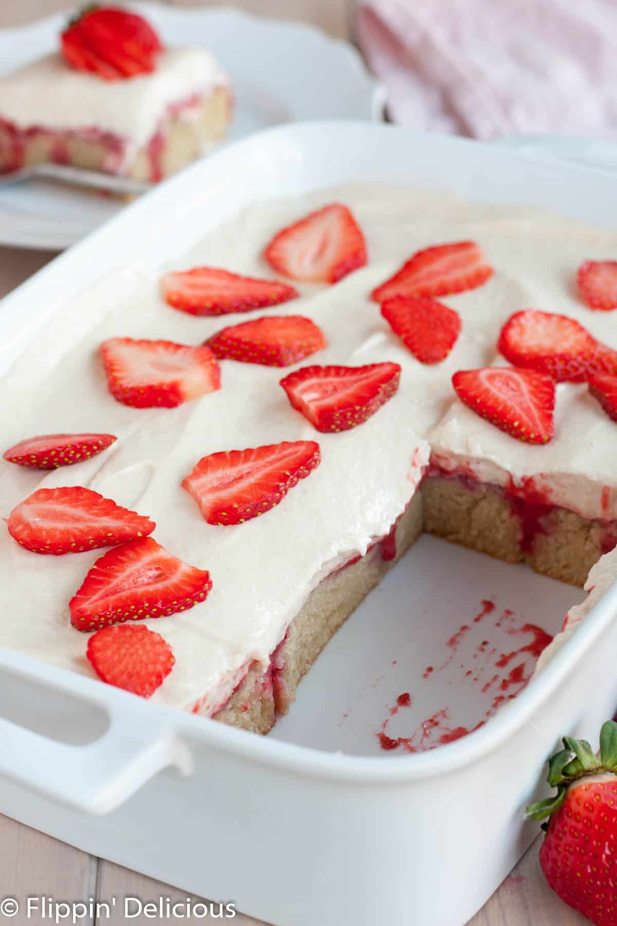 white ceramic rectangle pan filled with gluten free strawberries and cream poke cake , topped with sliced strawberries, with several slices removed