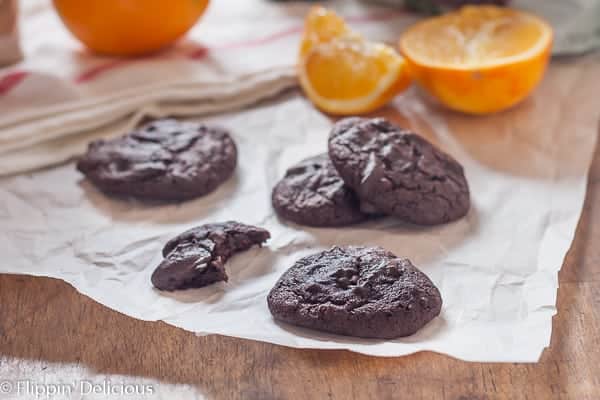 grain free coffee flour cookies, one with a bite taken out of it,  with cocoa powder and orange zest on a piece of parchment paper with slice oranges in the background with a red striped tea towel