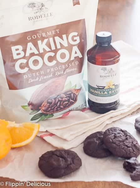 Rodelle baking cocoa and vanilla extract sitting beside grain free coffee flour cookies, one with a bite taken out of it,  with cocoa powder and orange zest on a piece of parchment paper with slice oranges in the background with a red striped tea towel