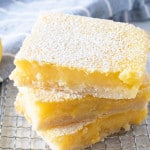 three gluten free lemon bars dusted with powdered sugar, stacked on top of each other on a wire cooling rack with a light blue striped dish towel in the background