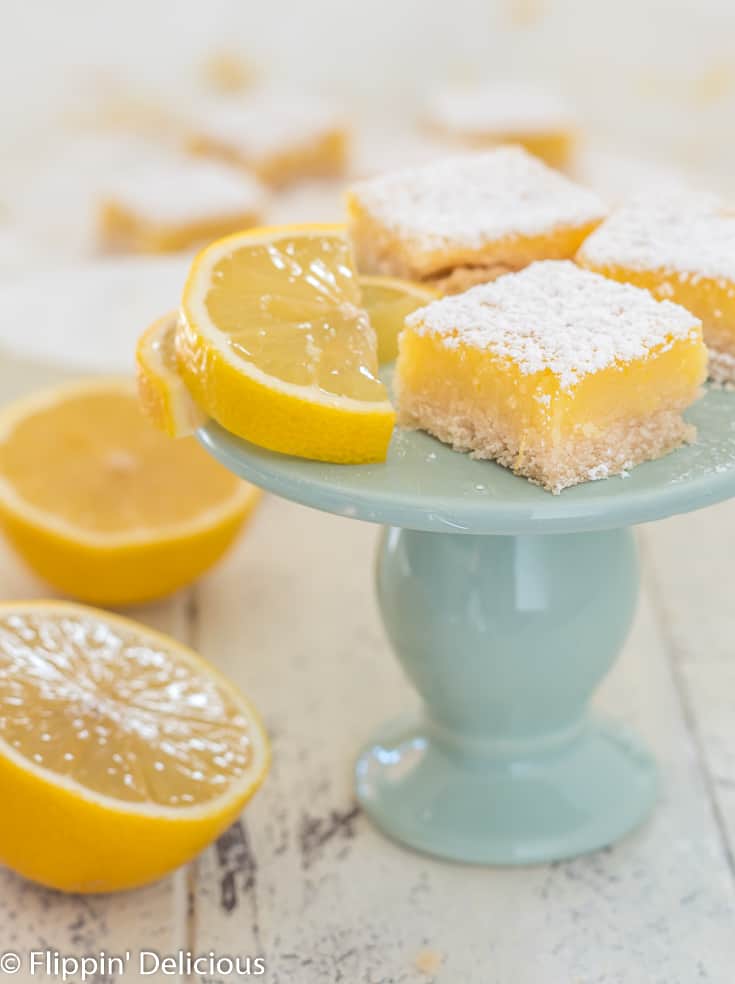 three gluten free lemon square bar cookies on small blue pedestal with lemon wedge and lemon cut in half
