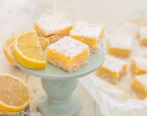 three gluten free lemon squares on blue plate next to lemon slice. More lemon squares and a lemon cut in half in the background