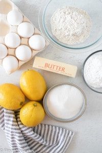 ingredients for gluten free lemon bars on a white table, whole lemons, eggs, a stick of butter, granulated sugar, powdered sugar, ad gluten free flour with a light blue striped dishtowel