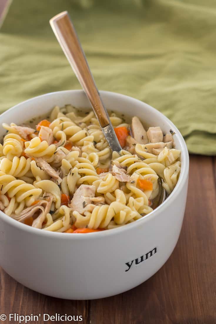 Instant Pot chicken noodle soup with gluten free pasta, sliced carrots, and herbes de Provence, in white bowl with wood handle spoon 