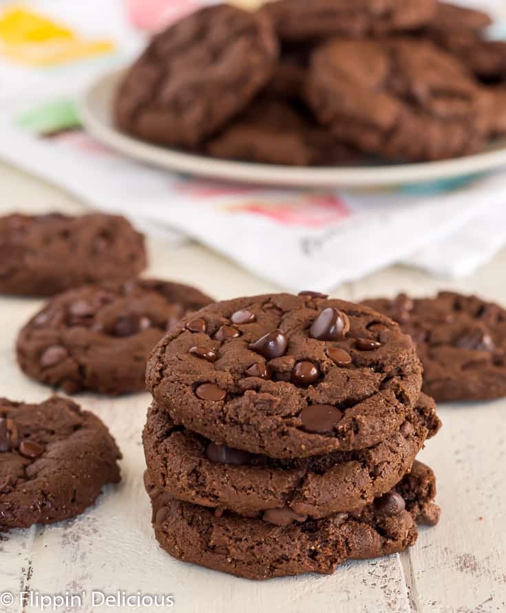 a stack with three gluten free dairy free double chocolate chip cookies on a white farm table with a plate of cookies in the back