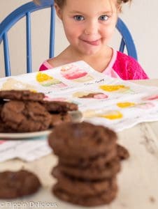 a girl in pink shirt and two side buns sticking out her tongue and drooling at gluten free double chocolate chip cookies in the foreground