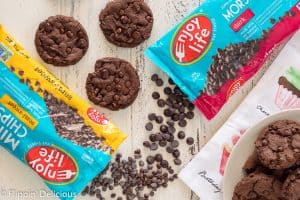 a bag of enjoy life mini chips and a bag of enjoy life dark morsels spilled onto a white farm table beside gluten free dairy free double chocolate chip cookies