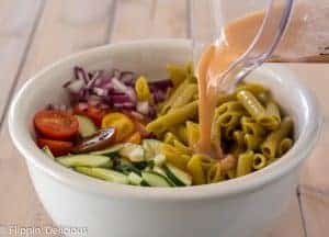 pouring homemade greek dressing from blender into large white bowl with gluten free green lentil penne, diced red onion, halved cherry tomatoes, and sliced cucumber